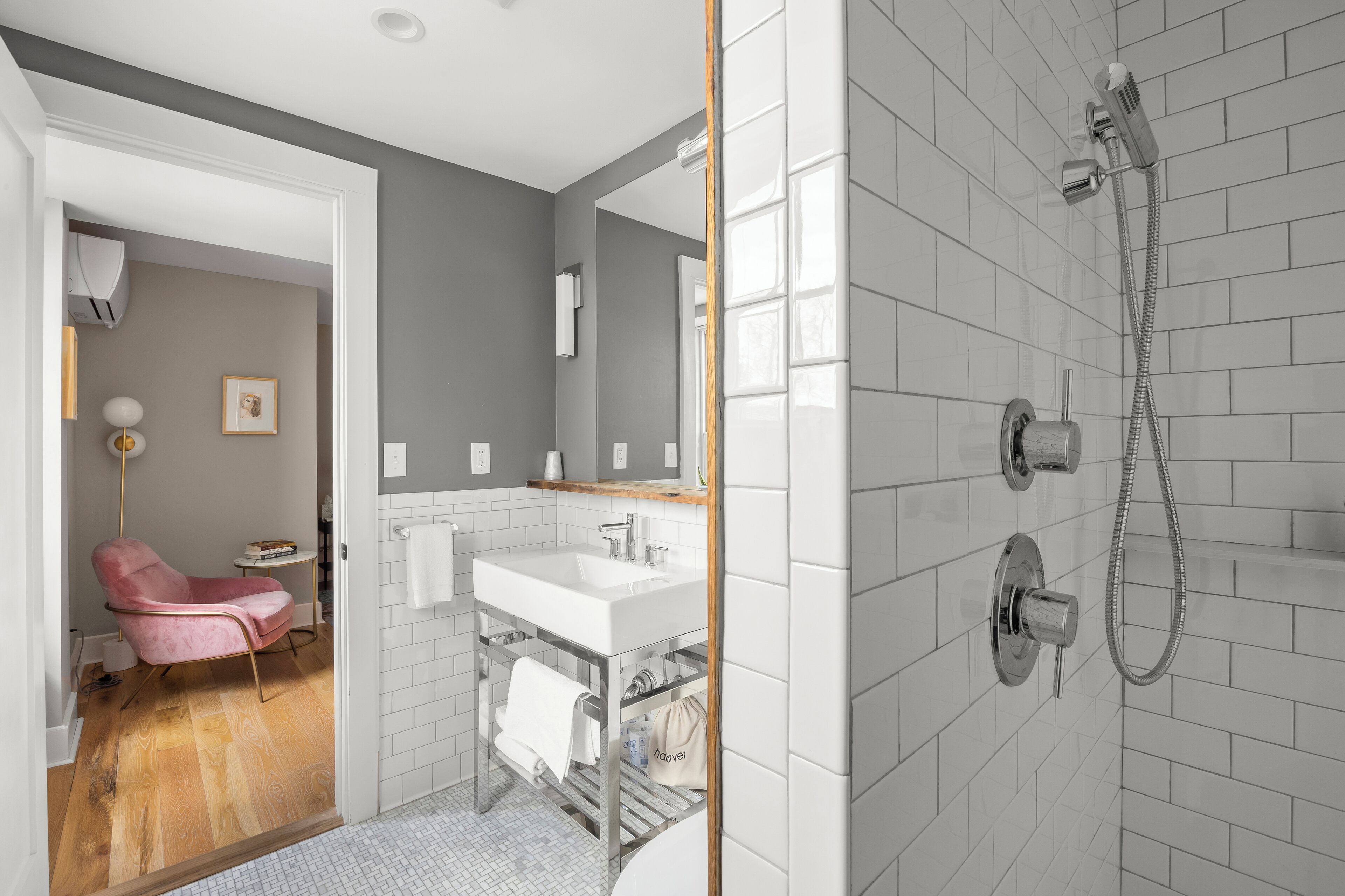 A modern bathroom with a shower, a sink, and a vanity. There is a chair in the adjacent room.