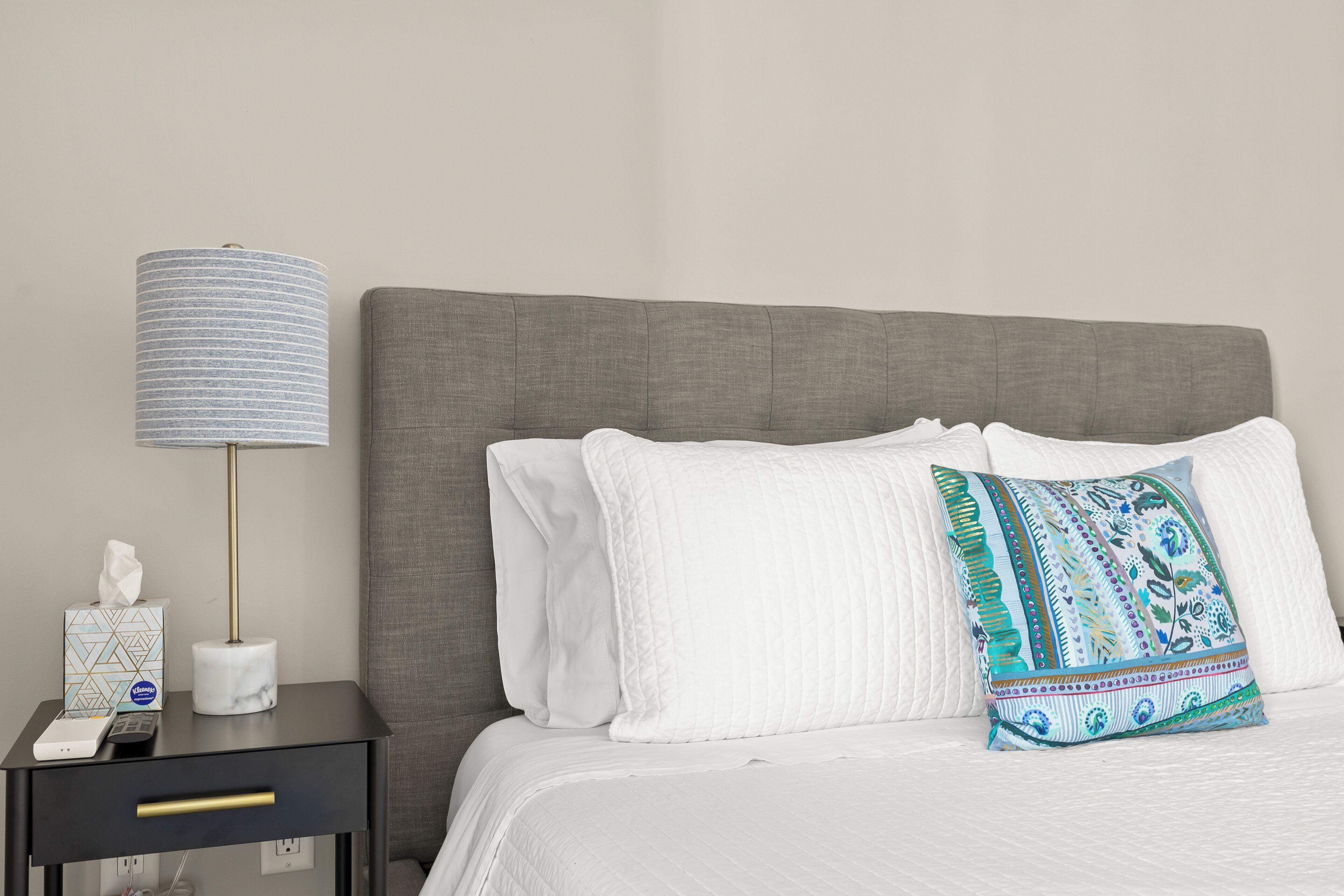 Un lit bien fait, avec une literie blanche et un coussin décoratif, une lampe de chevet et une table de chevet sur laquelle se trouvent une boîte de mouchoirs et une télécommande.