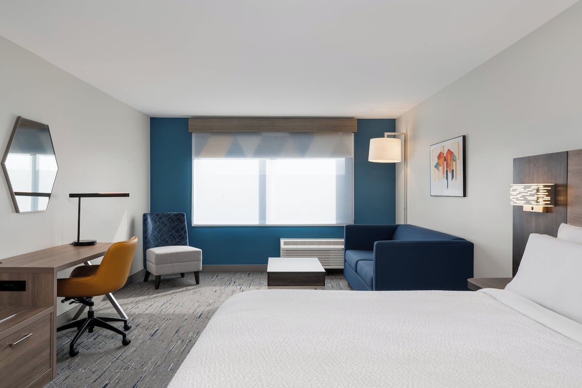 A hotel room with a bed, a desk, a chair, a sofa, a coffee table, and a window with blinds.