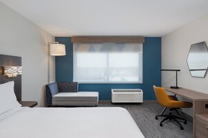 Habitación de hotel con cama, escritorio, silla, ventana con persianas, lámpara y espejo adosado a la pared.