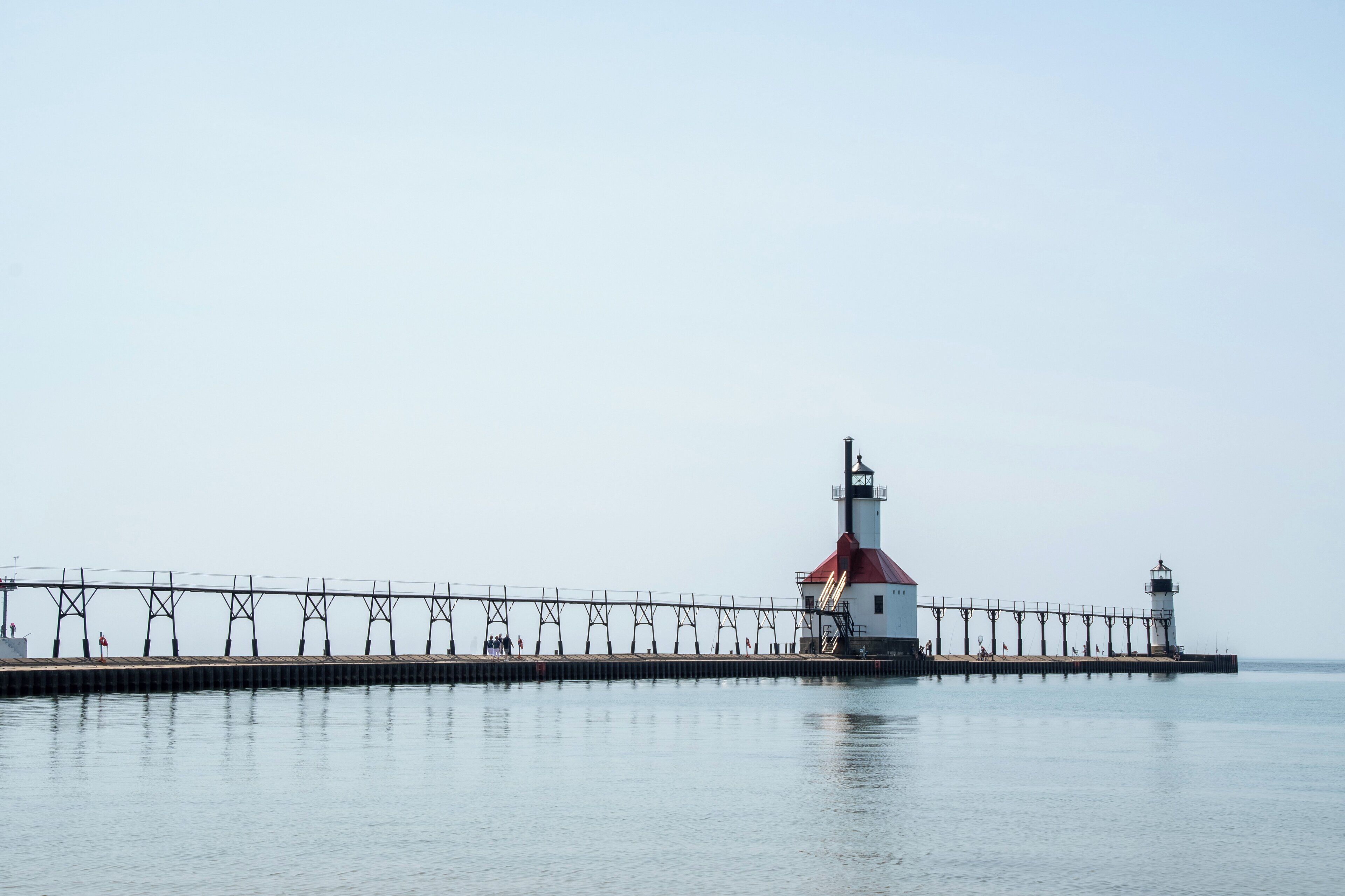 Un phare avec un toit rouge et un bâtiment blanc situé à l’extrémité d’une jetée qui s’avance dans une étendue d’eau calme.