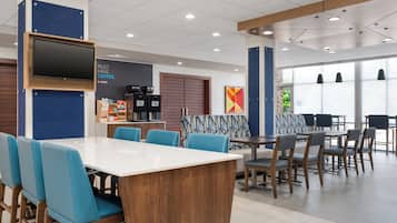 Un café moderno con una gran mesa blanca, sillas azules y una zona para preparar café.