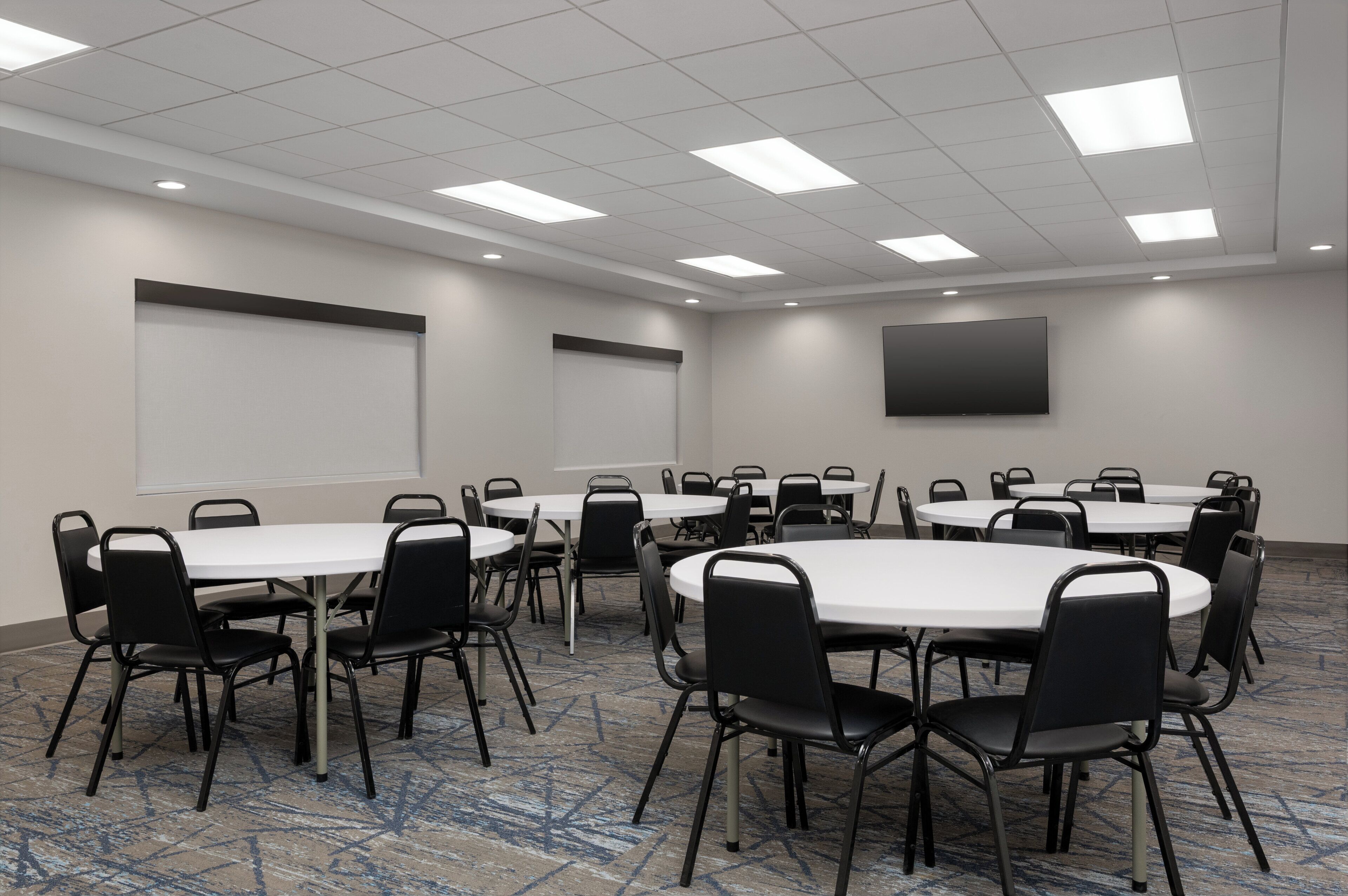 A conference room with multiple round tables and black chairs, a large flat-screen TV on the wall, and a carpeted floor.