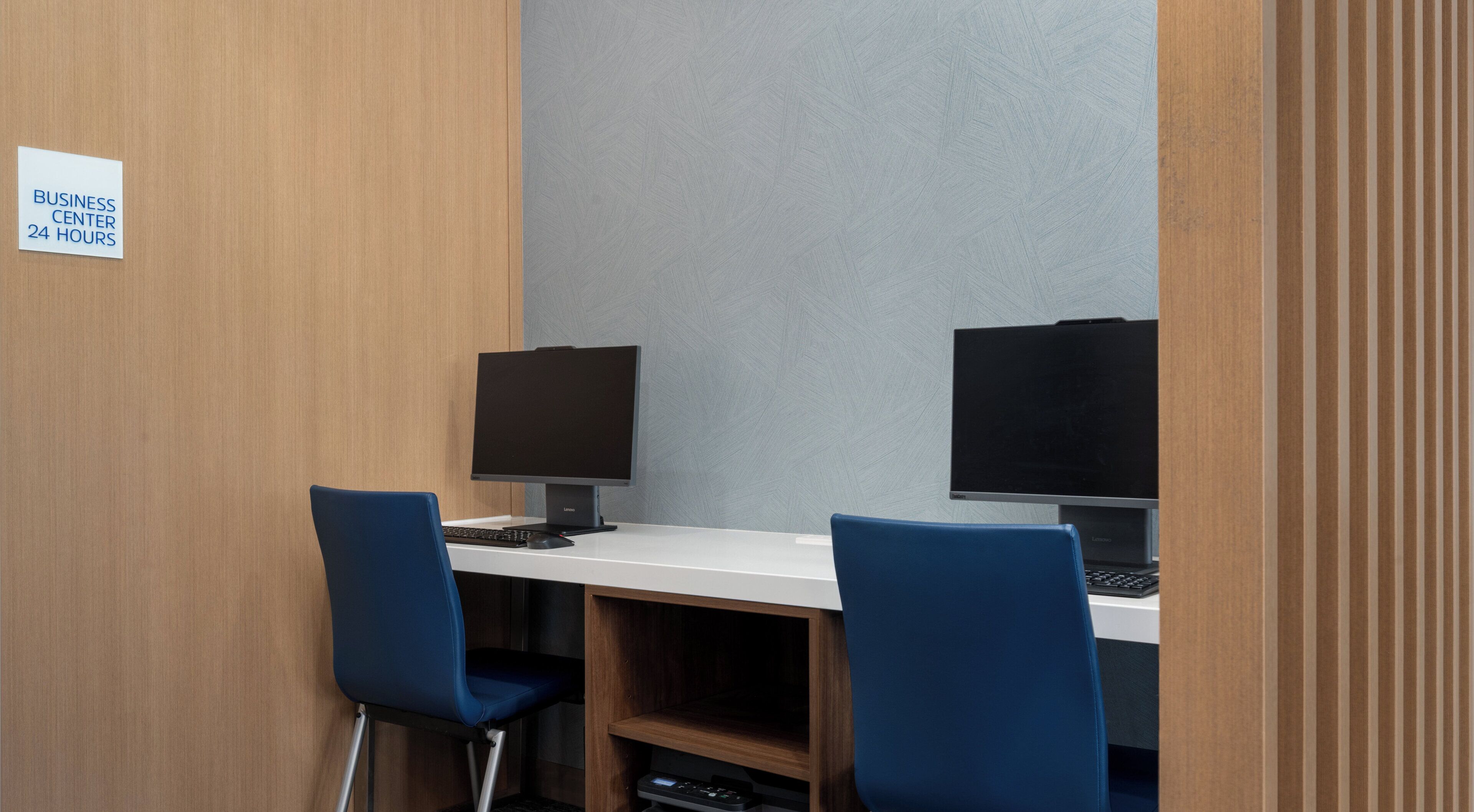 Un centro de negocios con dos estaciones de trabajo, cada una con un monitor y teclado, y dos sillas azules. Un letrero indica que el centro de negocios está abierto las 24 horas.