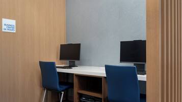 Un centro de negocios con dos estaciones de trabajo, cada una con un monitor y teclado, y dos sillas azules. Un letrero indica que el centro de negocios está abierto las 24 horas.