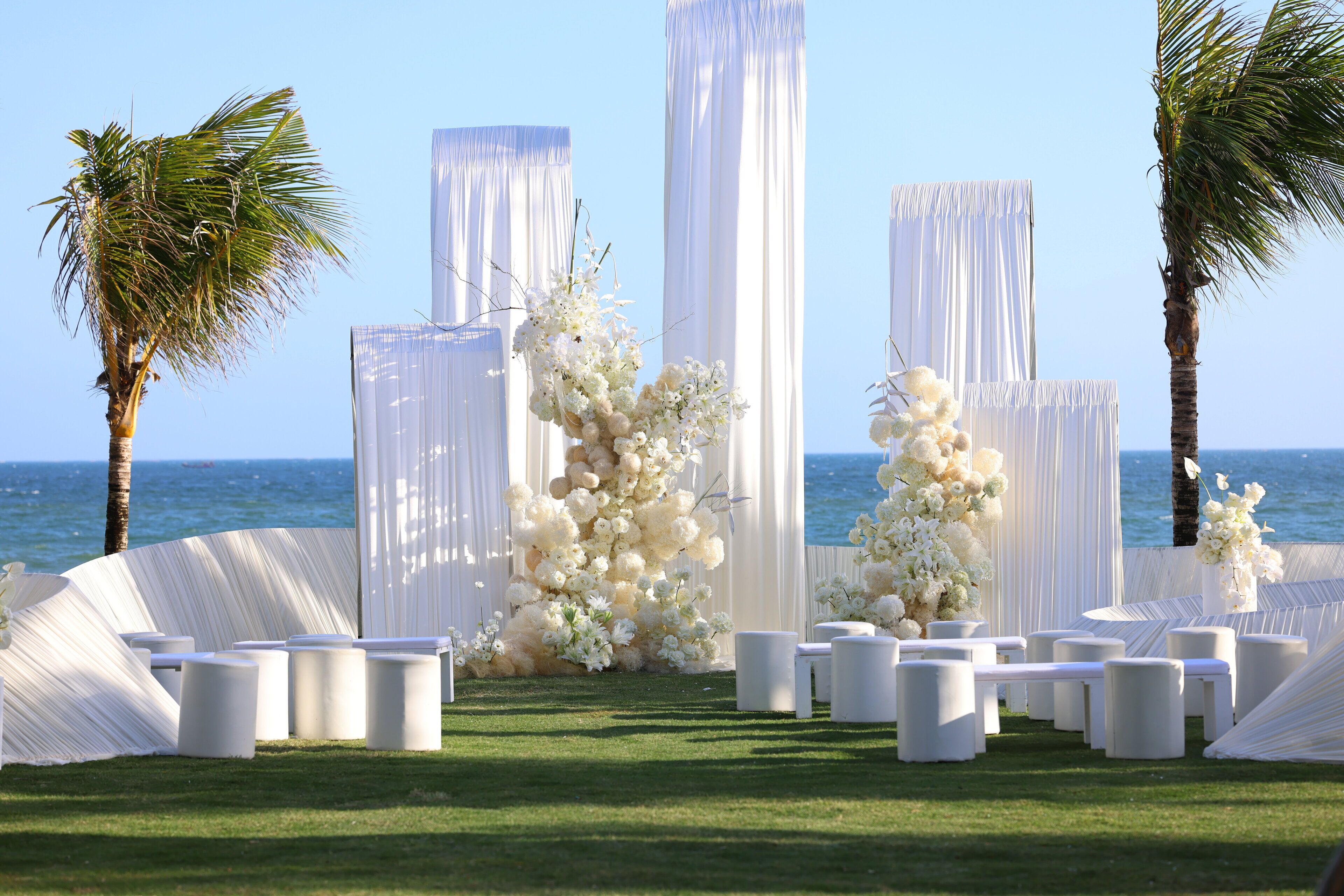 An outdoor wedding setup with white drapes, floral arrangements, and seating against a coastal backdrop.