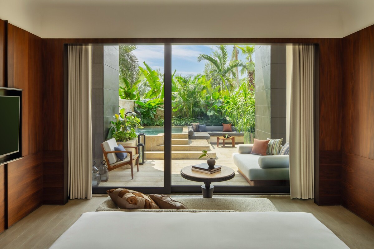 A modern hotel room with a large sliding glass door leading to a balcony with outdoor seating and a pool.