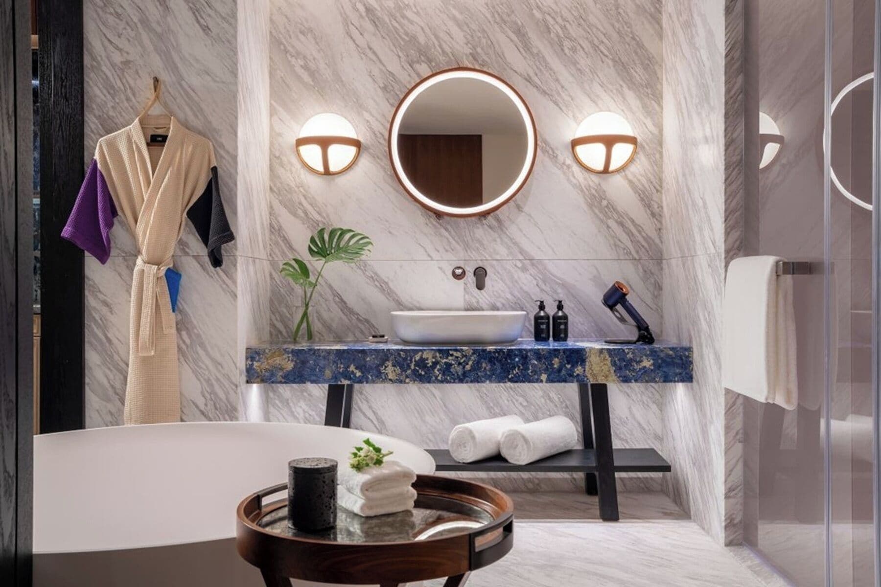 A modern bathroom with a marble wall, a round mirror, a sink with a blue countertop, and a bathtub.