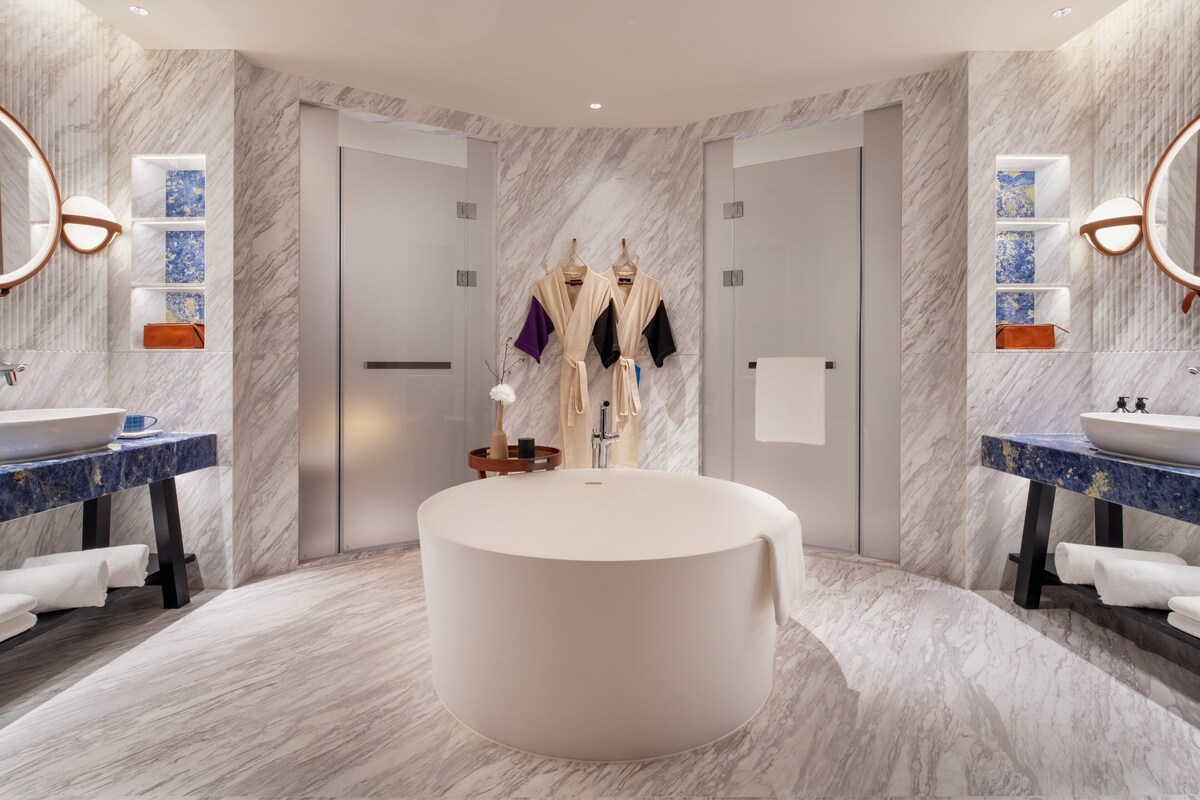 A modern bathroom with a central bathtub, two sinks, marble walls, and a mirror with wall sconces.