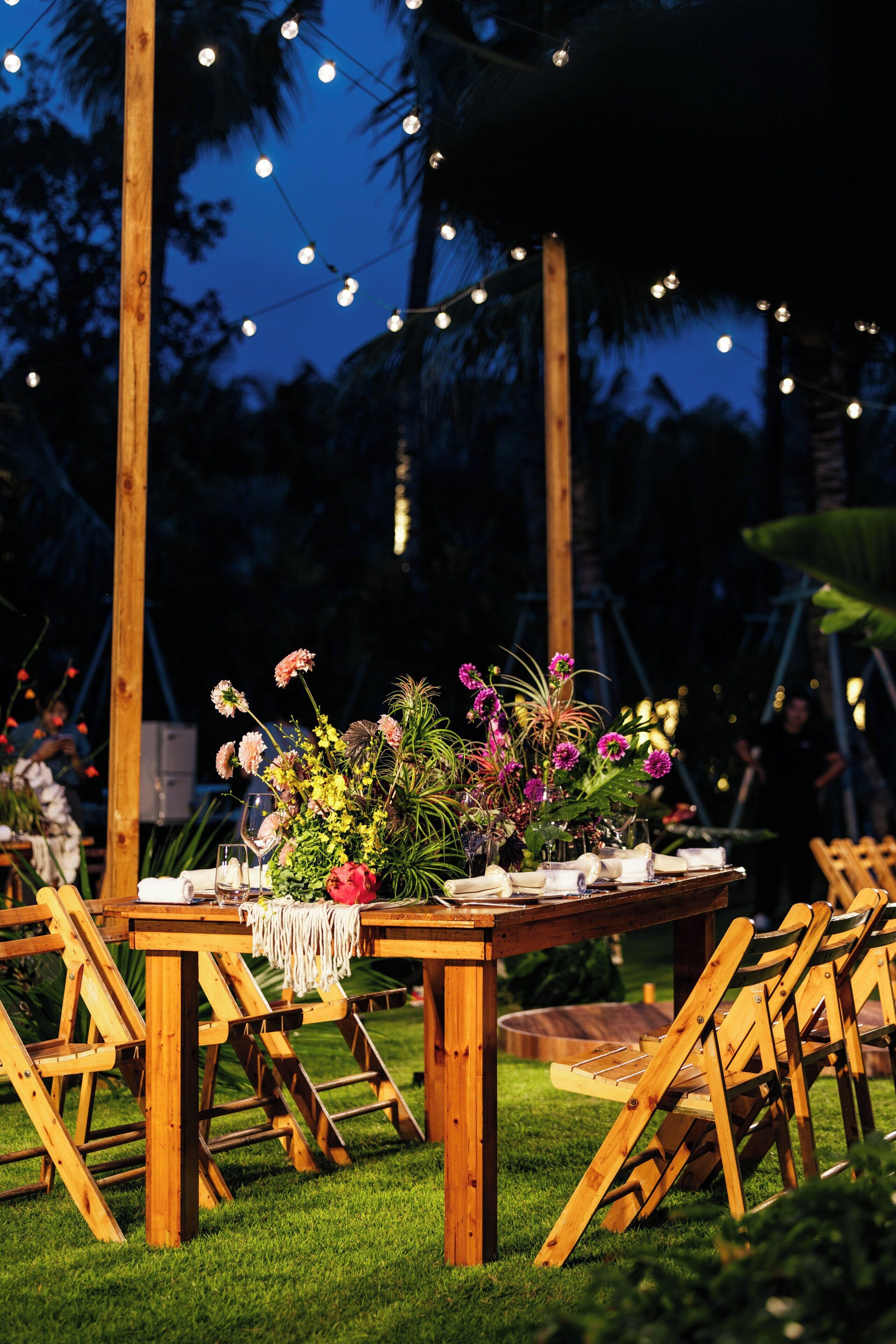 An outdoor wooden table with chairs, surrounded by greenery and string lights.