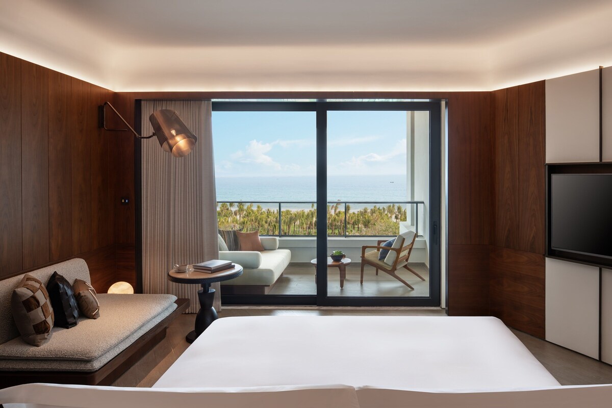 A modern hotel room with a large bed, a sofa, a TV, and a balcony with a view of the sea.