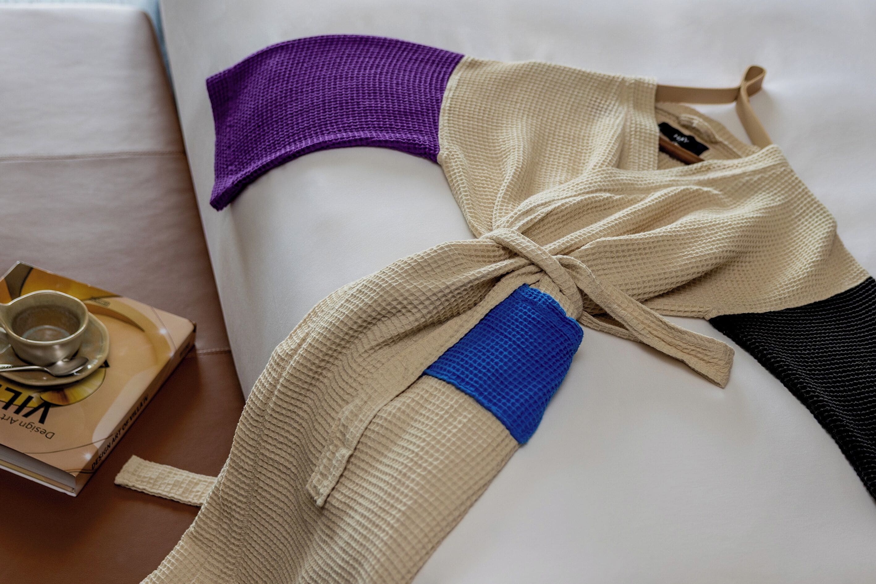 A bed with a beige and purple blanket, a beige and black sweater, and a book on a bedside table.