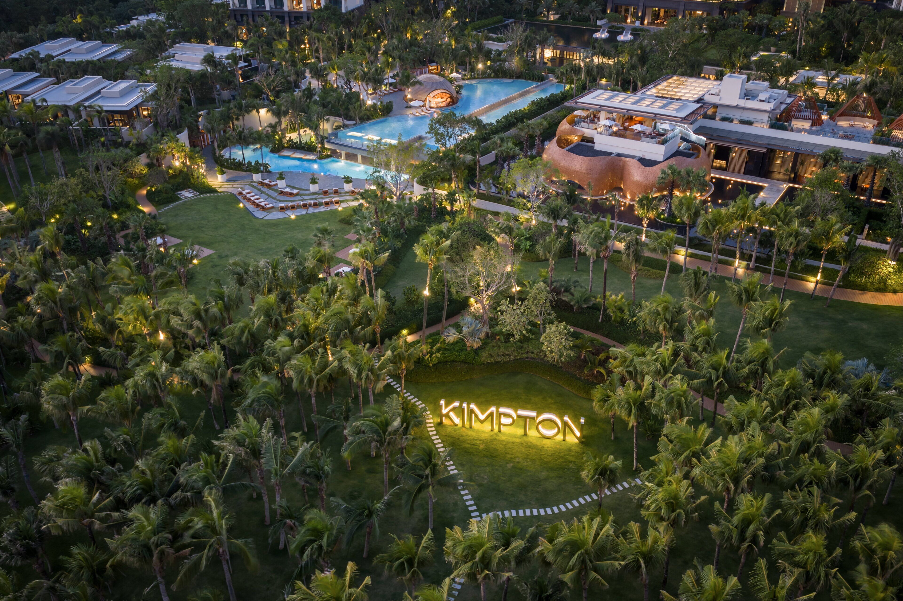 Een luchtbeeld van een resort met verschillende zwembaden, weelderige begroeiing en de naam 'KIMPTON' verlicht op de grond.