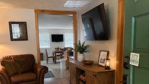 Un salón con un sofá de cuero marrón, suelo de madera, un televisor montado en la pared, una zona de comedor con mesa y sillas, y una puerta verde con un cartel.