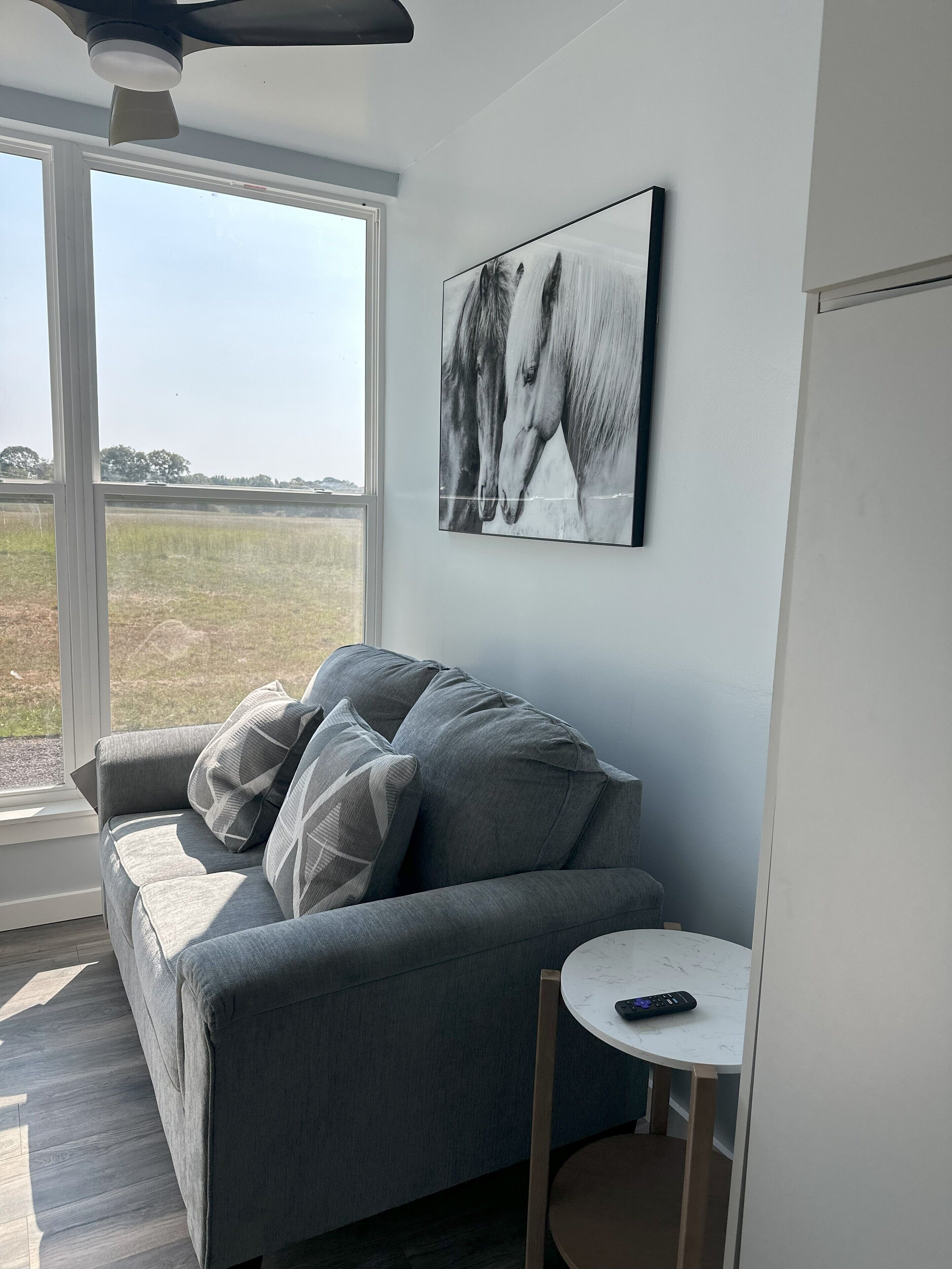 A modern living room with a grey sofa, a small round table, and a large window offering a view of a field.