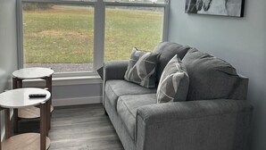 A room with a grey sofa, a ceiling fan, a large window with a view of a field, and a mounted picture of horses. - Magnolia Estate Container Home (Sardis)
