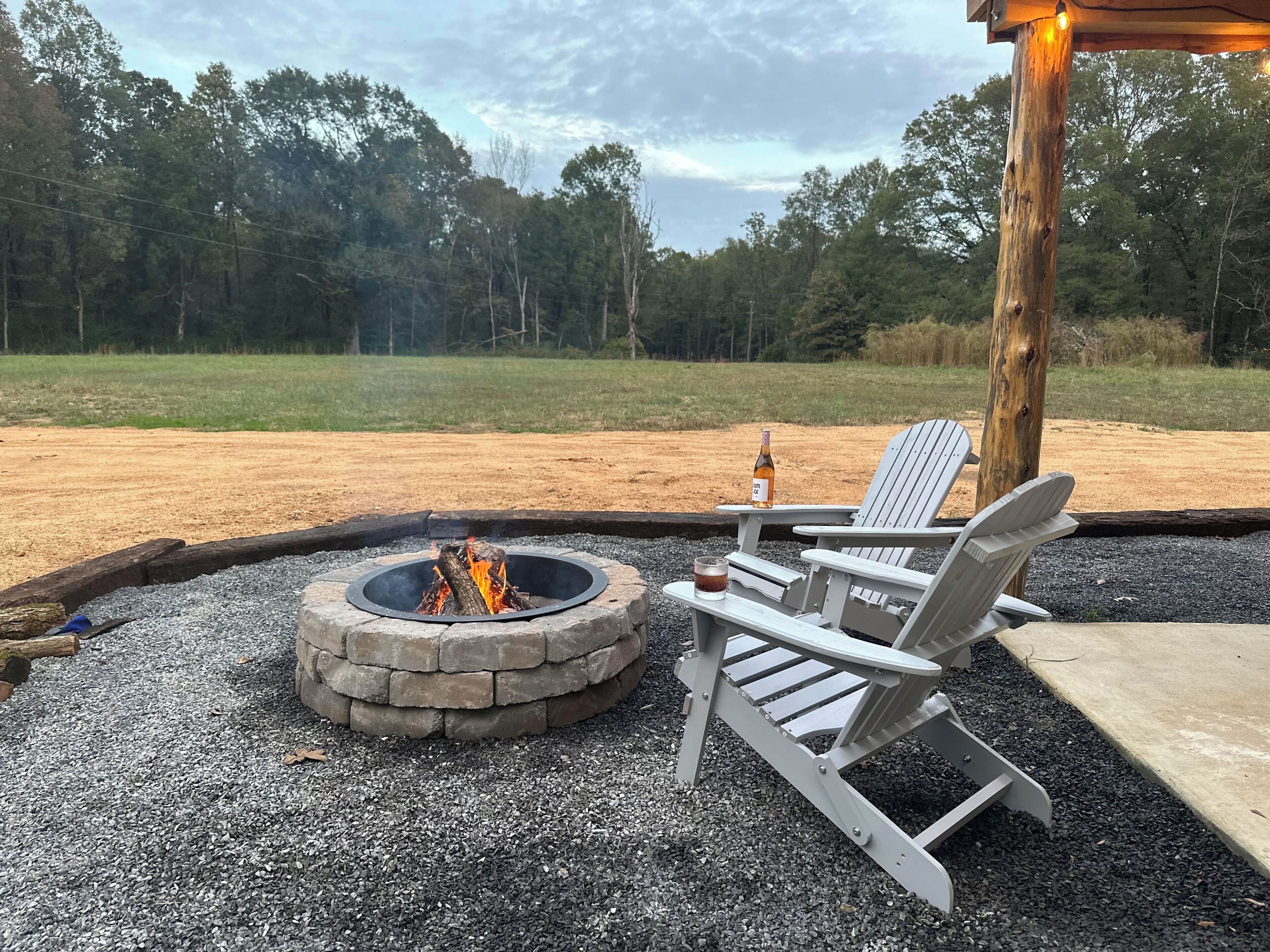 A fire pit with two Adirondack chairs and a bottle of wine, surrounded by gravel and a wooden deck.