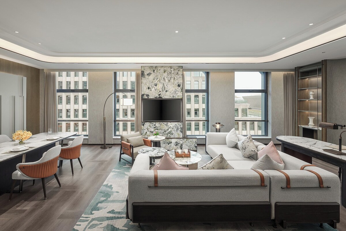 A modern living room with a sofa, dining table, and chairs, a TV mounted on the wall, and large windows offering views of the city.