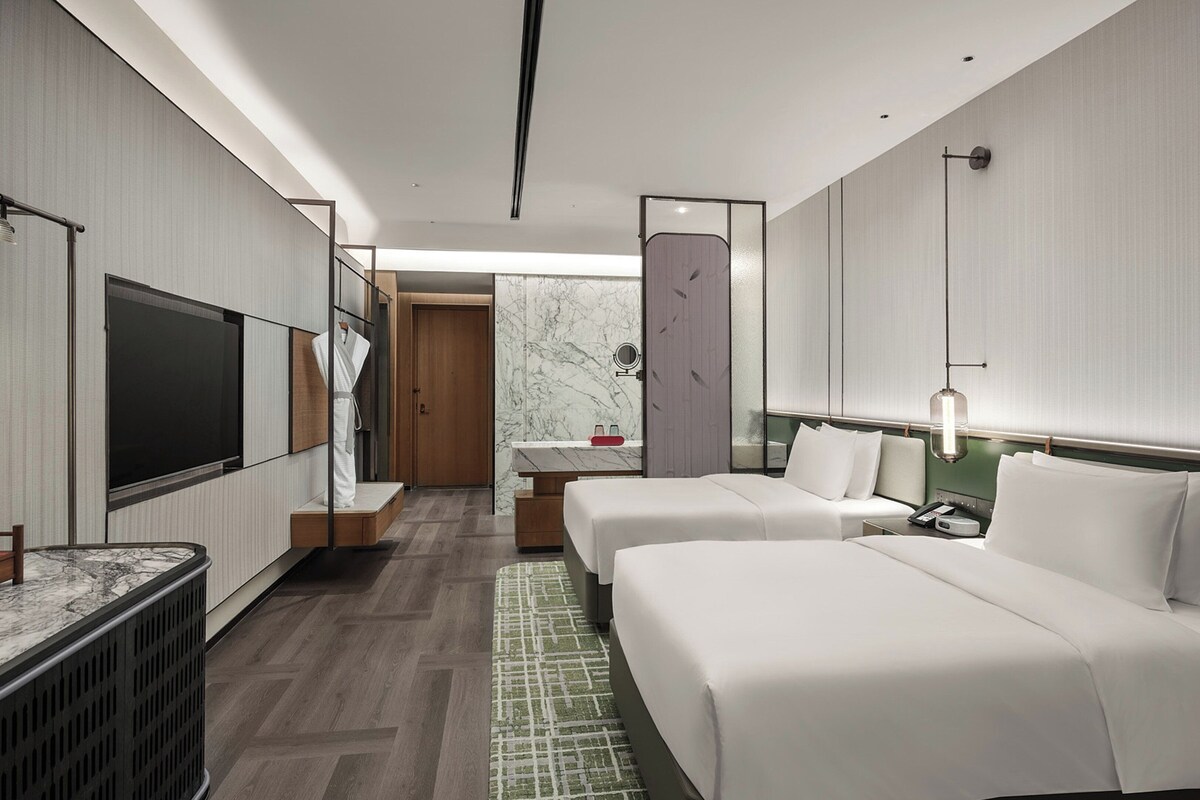 A hotel room with a TV, a bed, a bedside table, a mirror, and a bathroom with a marble wall.
