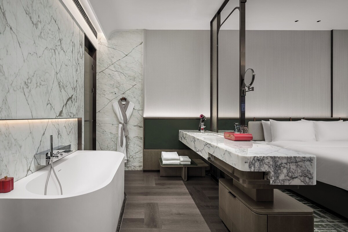 A modern bathroom with a marble wall, a freestanding bathtub, a vanity with a mirror, and a bed with a headboard.