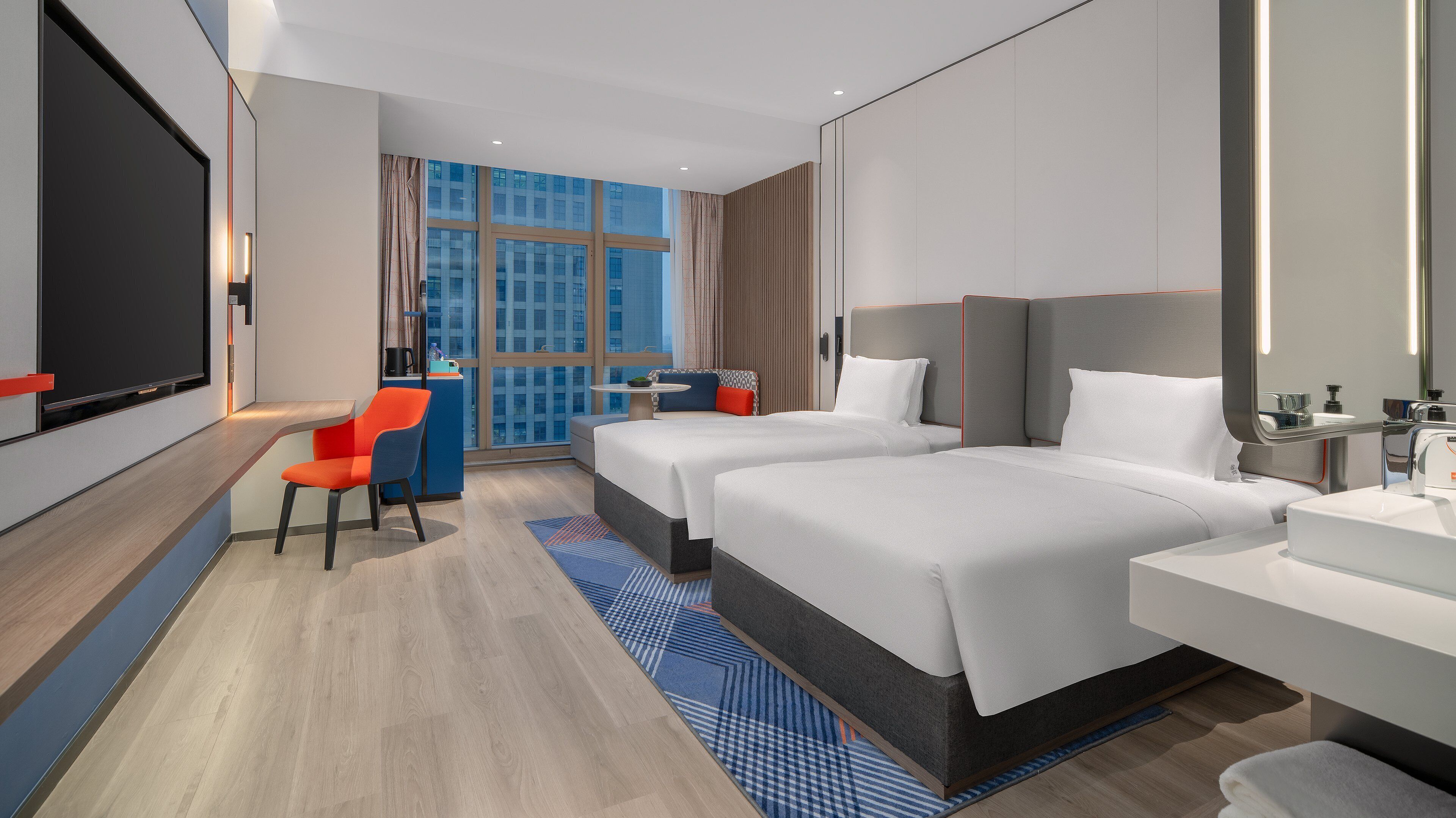 A hotel room with two beds, a desk, a chair, a large window, and a flat-screen TV.