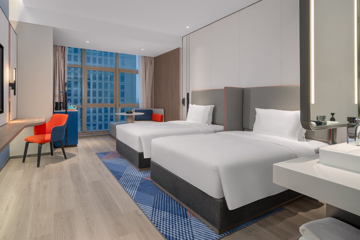 A hotel room with two beds, a desk, a chair, a large window, and a flat-screen TV.