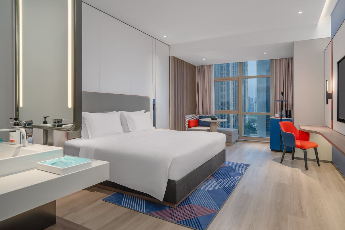 A modern hotel room with a large bed, a desk, a chair, a TV, and a view of the city.