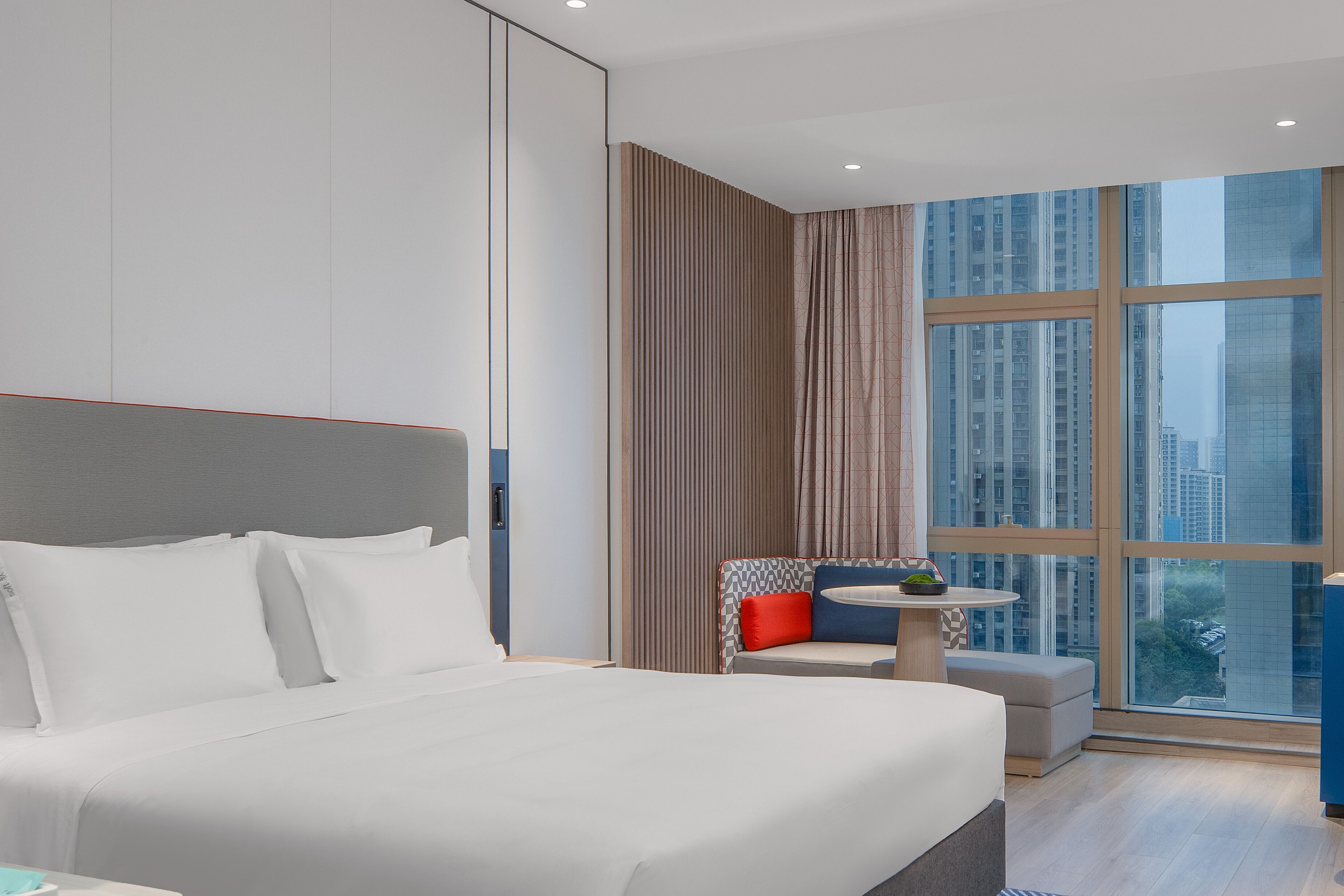 A modern hotel room with a large bed, a small seating area, and a large window offering a city view.
