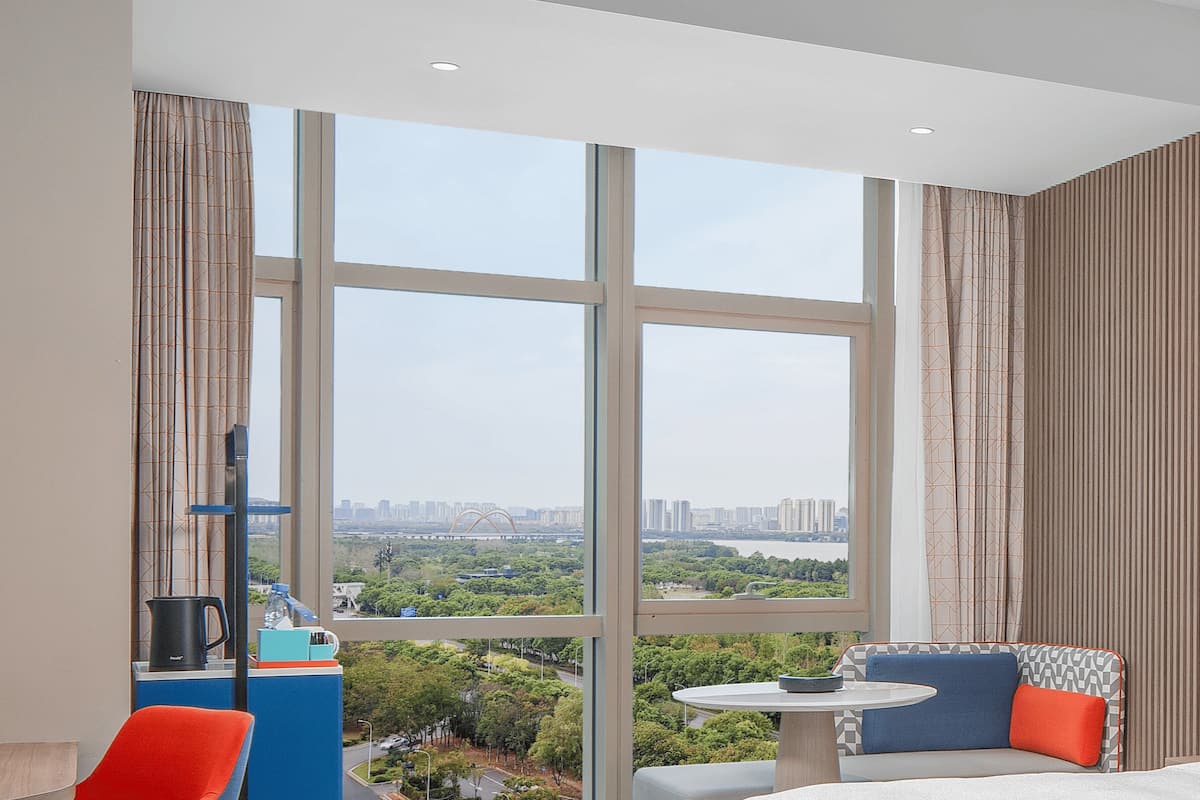 A modern hotel room with a large window offering a city view, a small round table, and a sofa with colorful cushions.