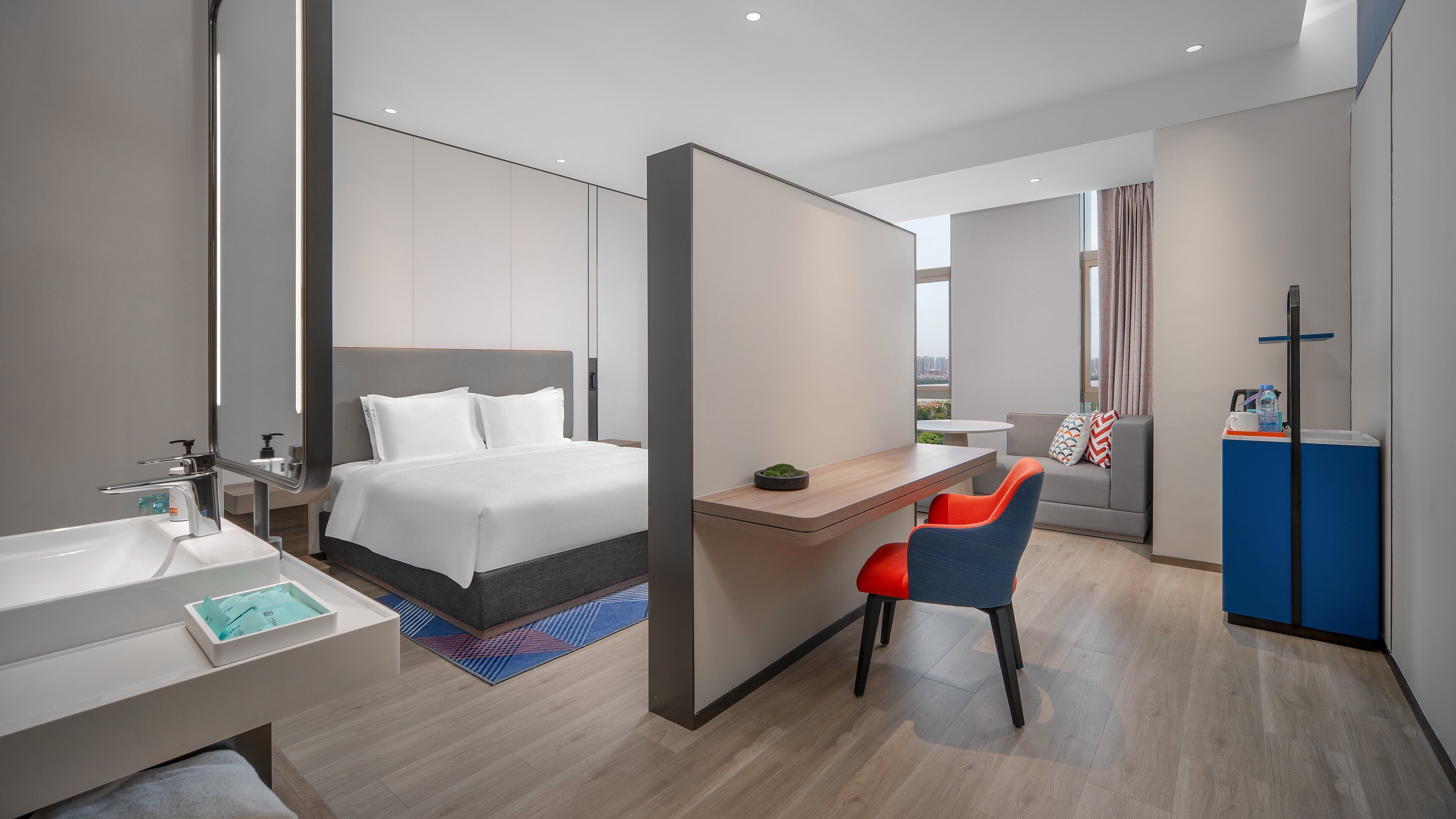 A modern hotel room with a large bed, a desk, a chair, a small table, a sink, and a mirror.