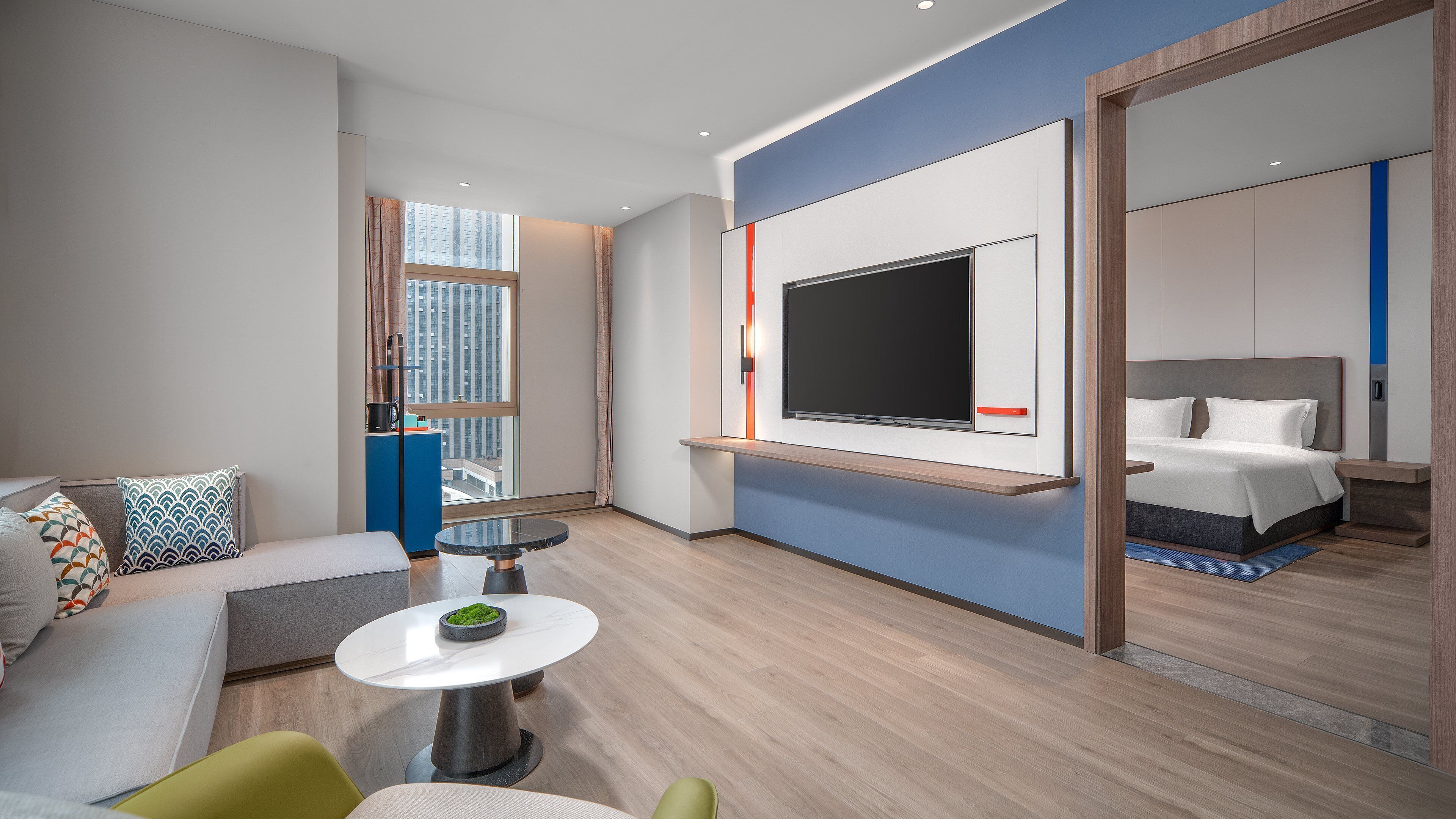 Moderni hotellihuone, jossa on suuri sänky, pieni pöytä ja oleskelualue sekä seinälle asennettu televisio.
