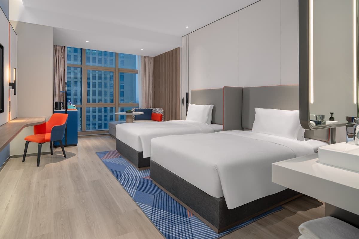 A hotel room with two beds, a desk, a chair, a large window, and a flat-screen TV.