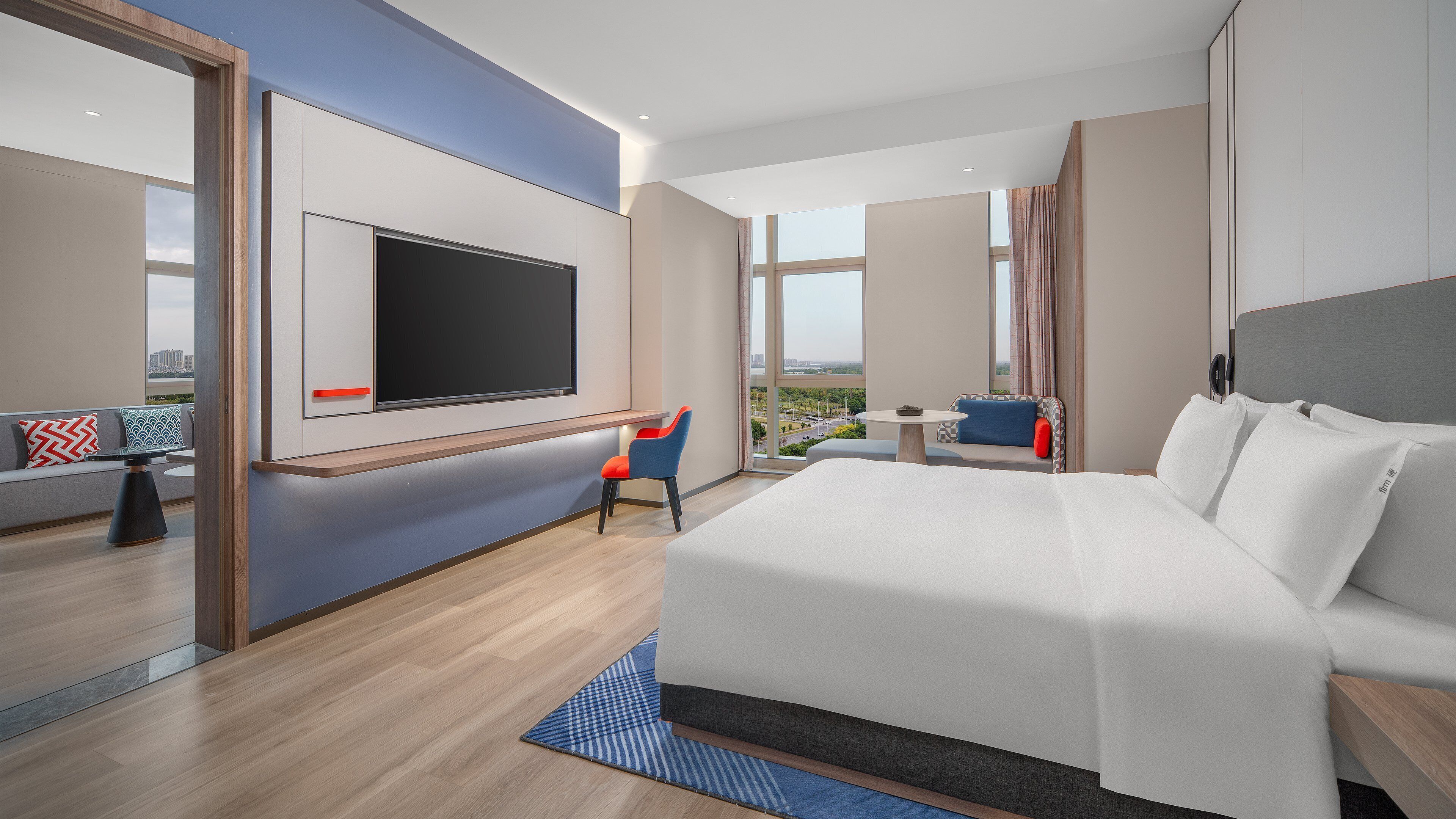 Una habitación de hotel moderna con una cama grande, un televisor montado en la pared, una zona de estar con una mesa y ventanas amplias con vistas a la ciudad.