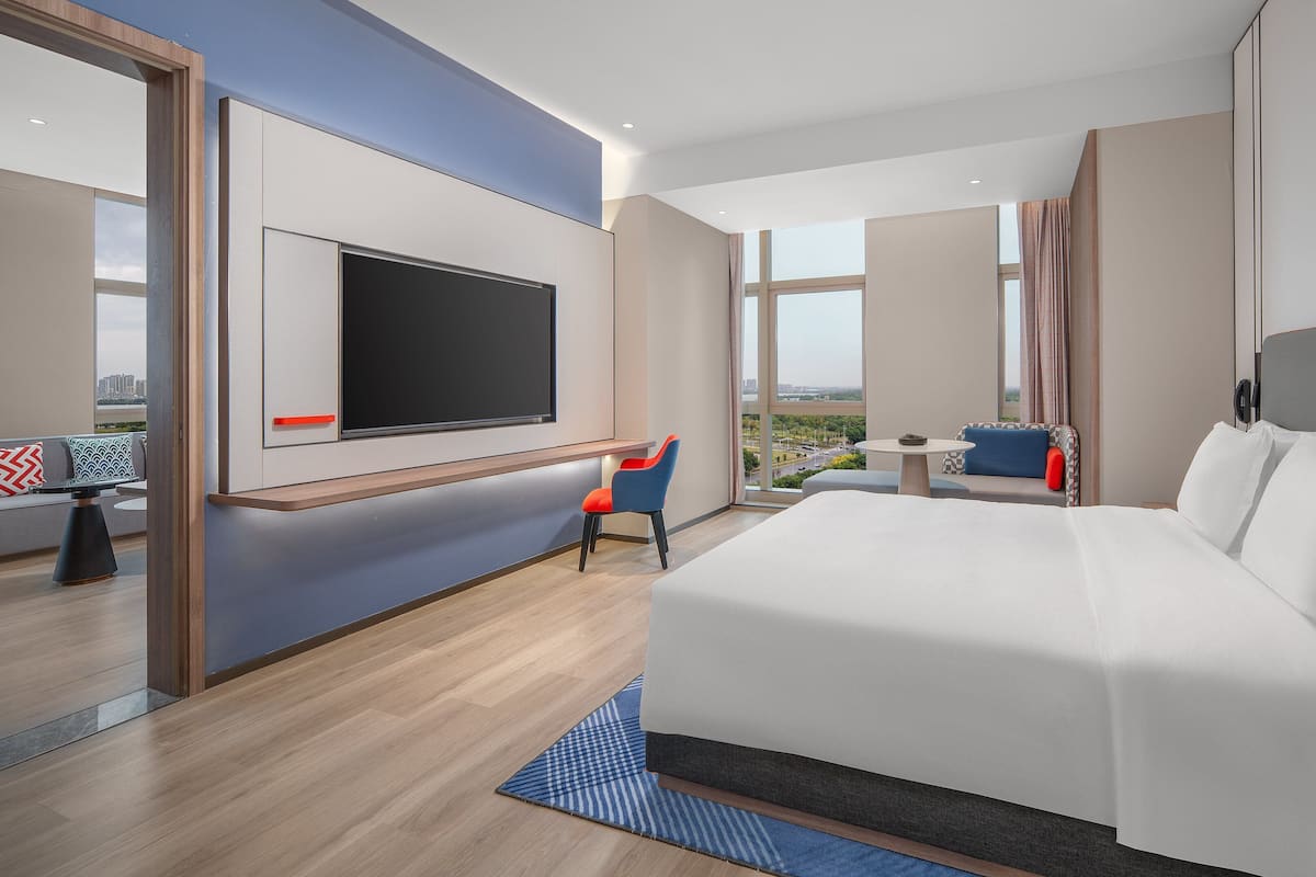 A modern hotel room with a large bed, a TV mounted on the wall, a seating area with a table, and large windows offering a city view.
