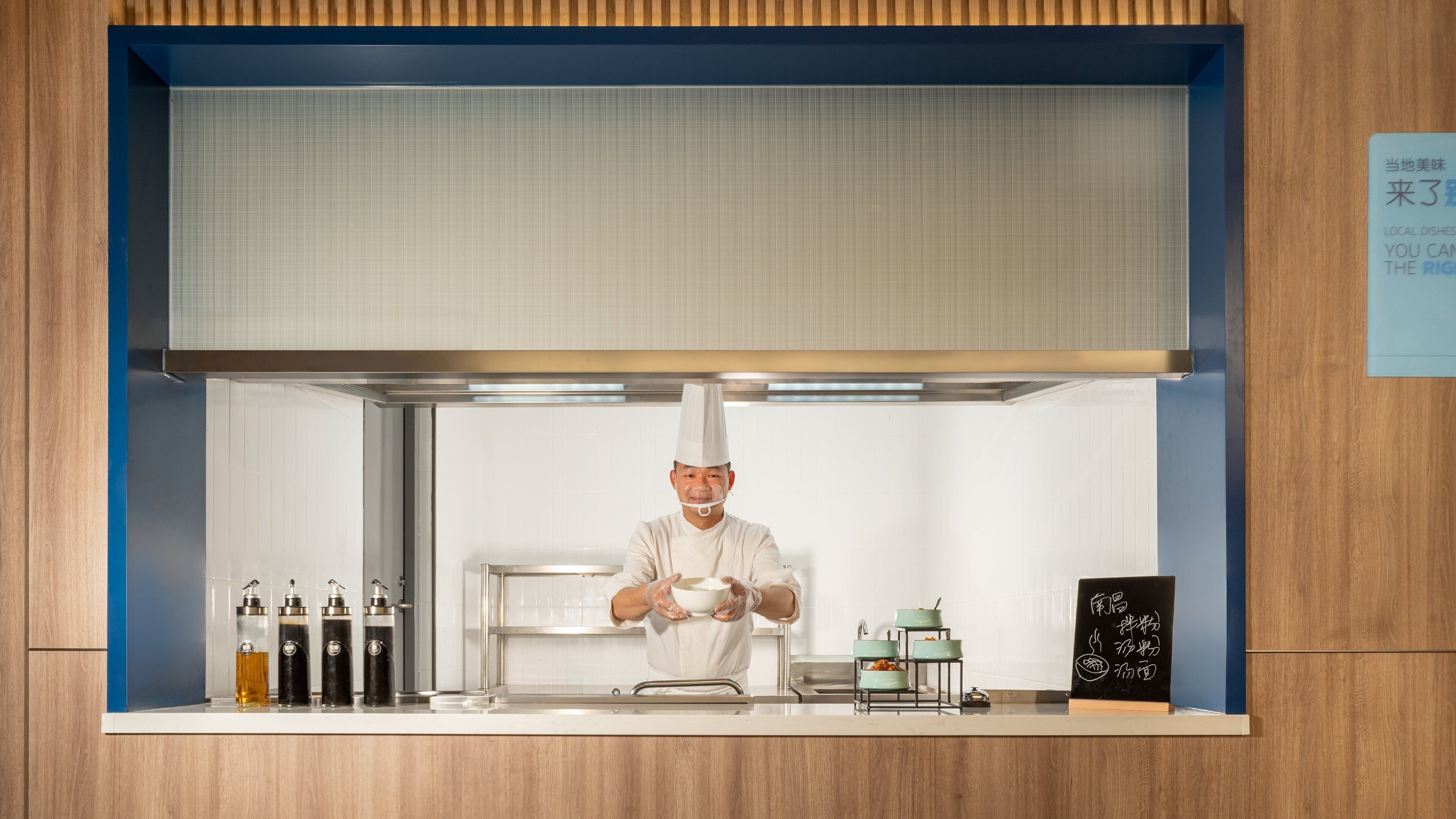 Un chef en una cocina con una decoración azul y blanca, electrodomésticos de acero inoxidable y un menú en una pizarra.
