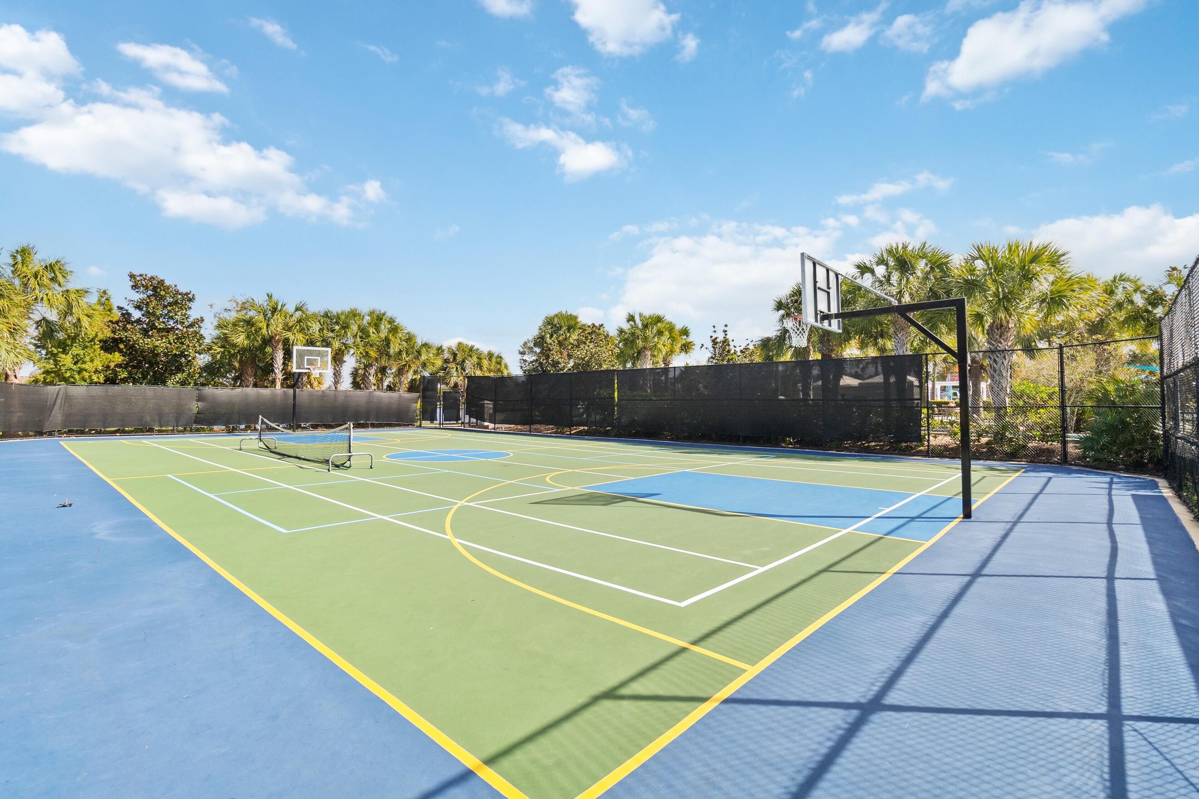 An outdoor basketball court with a clear sky, palm trees, and a fence.