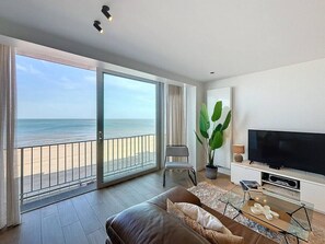 A modern living room with a large sliding glass door leading to a balcony with a view of the beach.