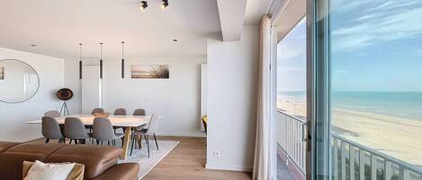 Un salon moderne doté d’une grande fenêtre donnant sur une plage, un coin repas avec une table ronde et des chaises, et un canapé en cuir marron.