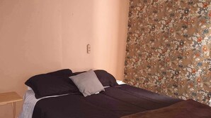 Una cama individual con una colcha oscura y almohadas, una mesita de noche de madera y un cortin con estampado floral.