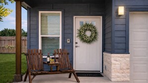 L’extérieur d’une maison avec une porte d’entrée, une guirlande, un banc en bois et une table sur laquelle sont posés divers objets.