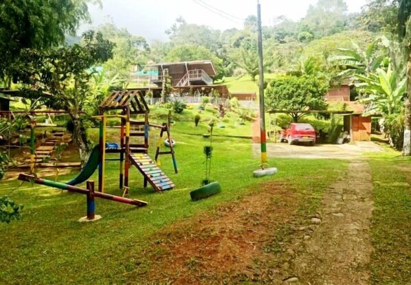 A playground with swings and a slide, surrounded by greenery and a house in the background.