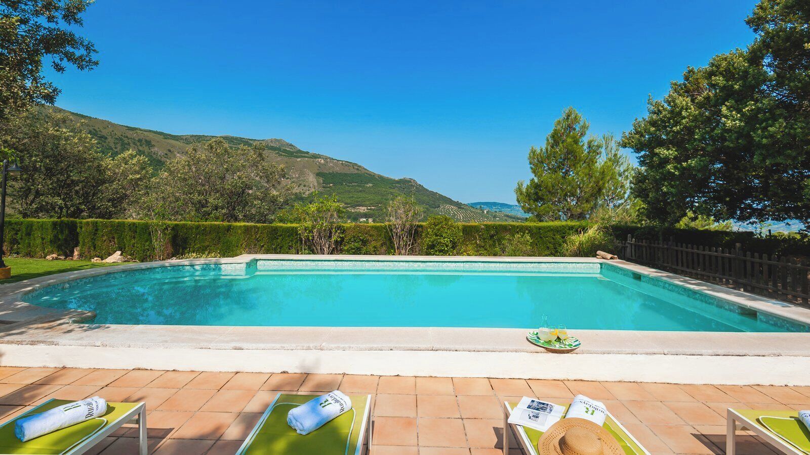 A rectangular swimming pool with a tiled deck, surrounded by a wooden fence and lush greenery.