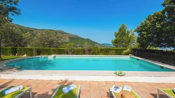 A rectangular swimming pool with a tiled deck, surrounded by a wooden fence and lush greenery.