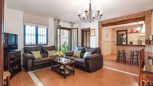 A living room with two sofas, a coffee table, and a TV. There is a kitchen area with stools and a brick wall.