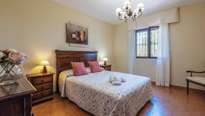 A bedroom with a bed, a nightstand, a lamp, a chair, a window with curtains, and a painting on the wall. - Azuara 3 Dormitorios Los Villares by Ruralidays (Jaén)