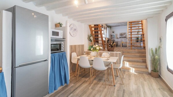 Een moderne keuken met een roestvrijstalen koelkast, een magnetron en een inbouwoven. Er is een eethoek met een tafel gedekt voor twee personen, witte stoelen en een vaas met bloemen. Een houten trap leidt naar een ander niveau.