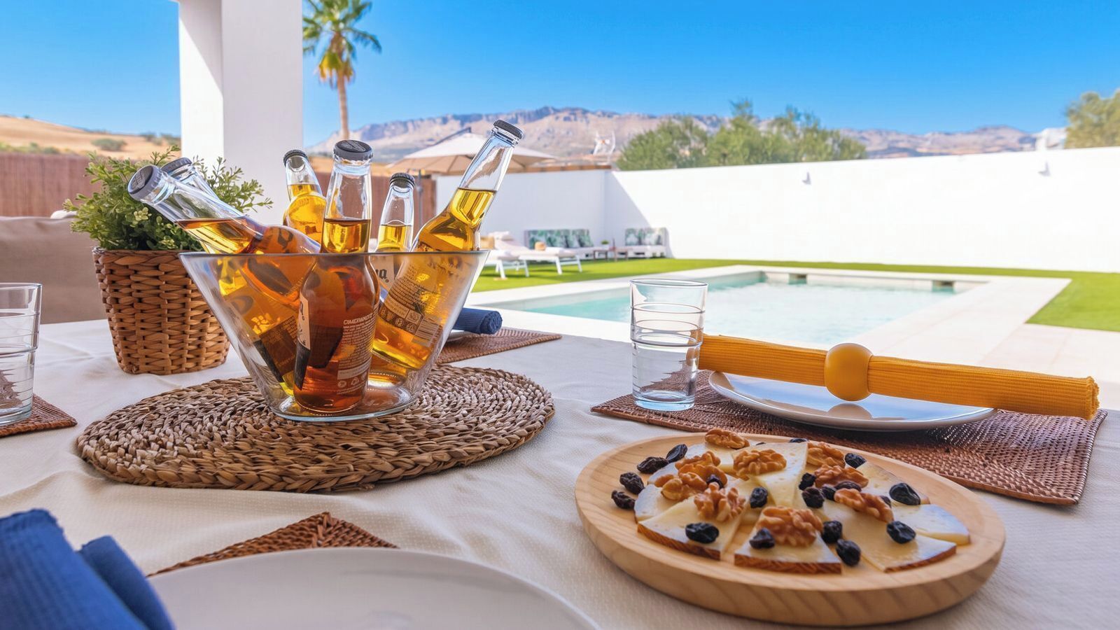 A table set with a bottle of wine, glasses, a wooden board with cheese and nuts, and a view of a pool and mountains.