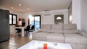 A living room with a sofa, a dining table with chairs, a kitchen area, and a view through a glass door.