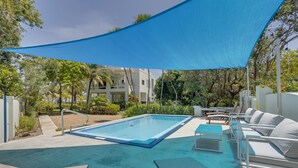Una piscina con sillas de descanso y una zona con sombra, rodeada de vegetación y edificios.