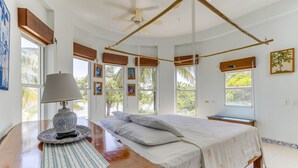 Un dormitorio con cama, una mesita de noche con lámpara y vistas a palmeras a través de amplias ventanas.